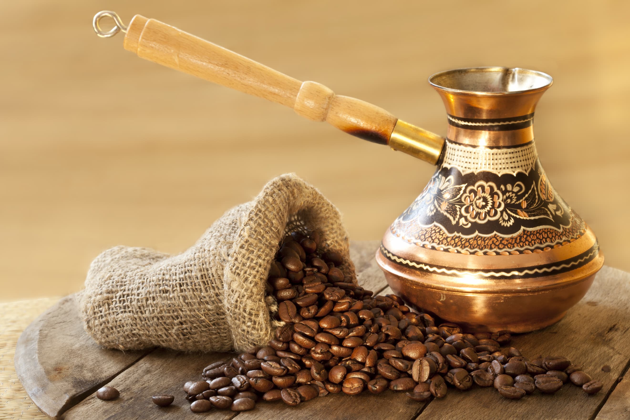 Aus einem Jutesack fallende Kaffeebohnen neben traditioneller Kupferkanne auf Holzbrett.