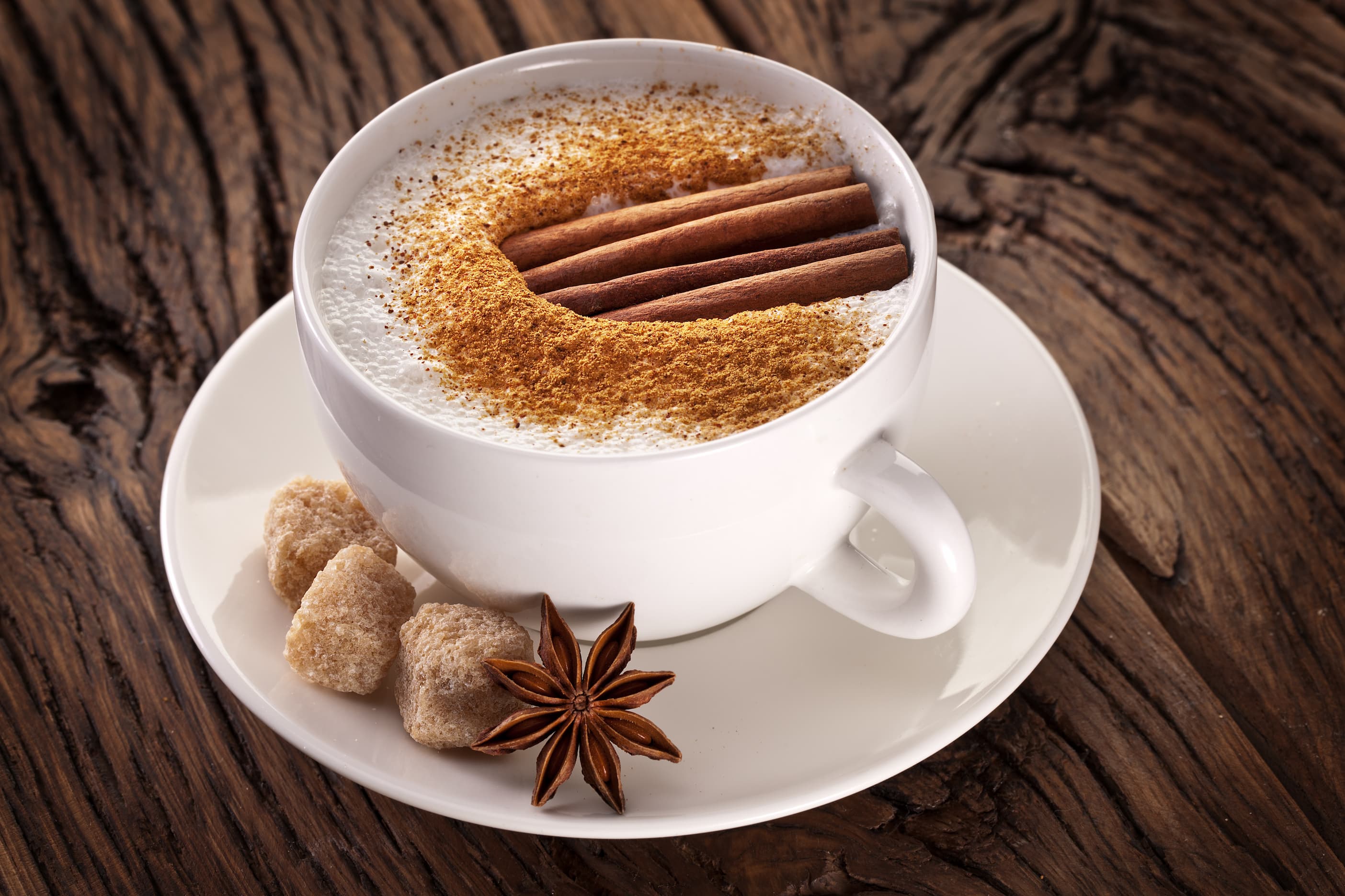 Cappuccino in weißer Tasse mit Milchschaum, Zimt und Zuckerwürfeln auf Holztisch.