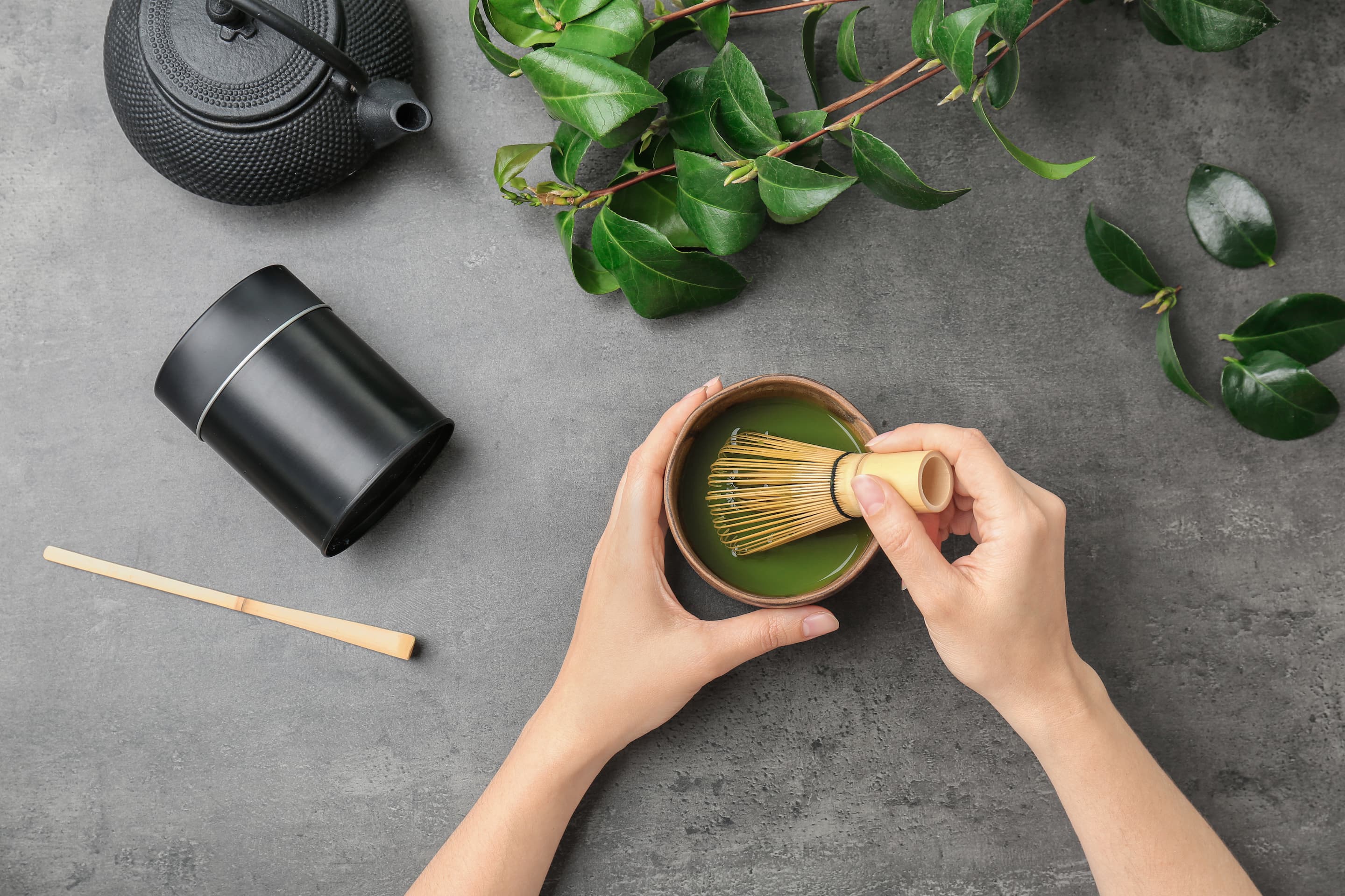 Hände bereiten Matcha Tee mit Bambusbesen in Schale zu, Teekanne und Pflanzen daneben.