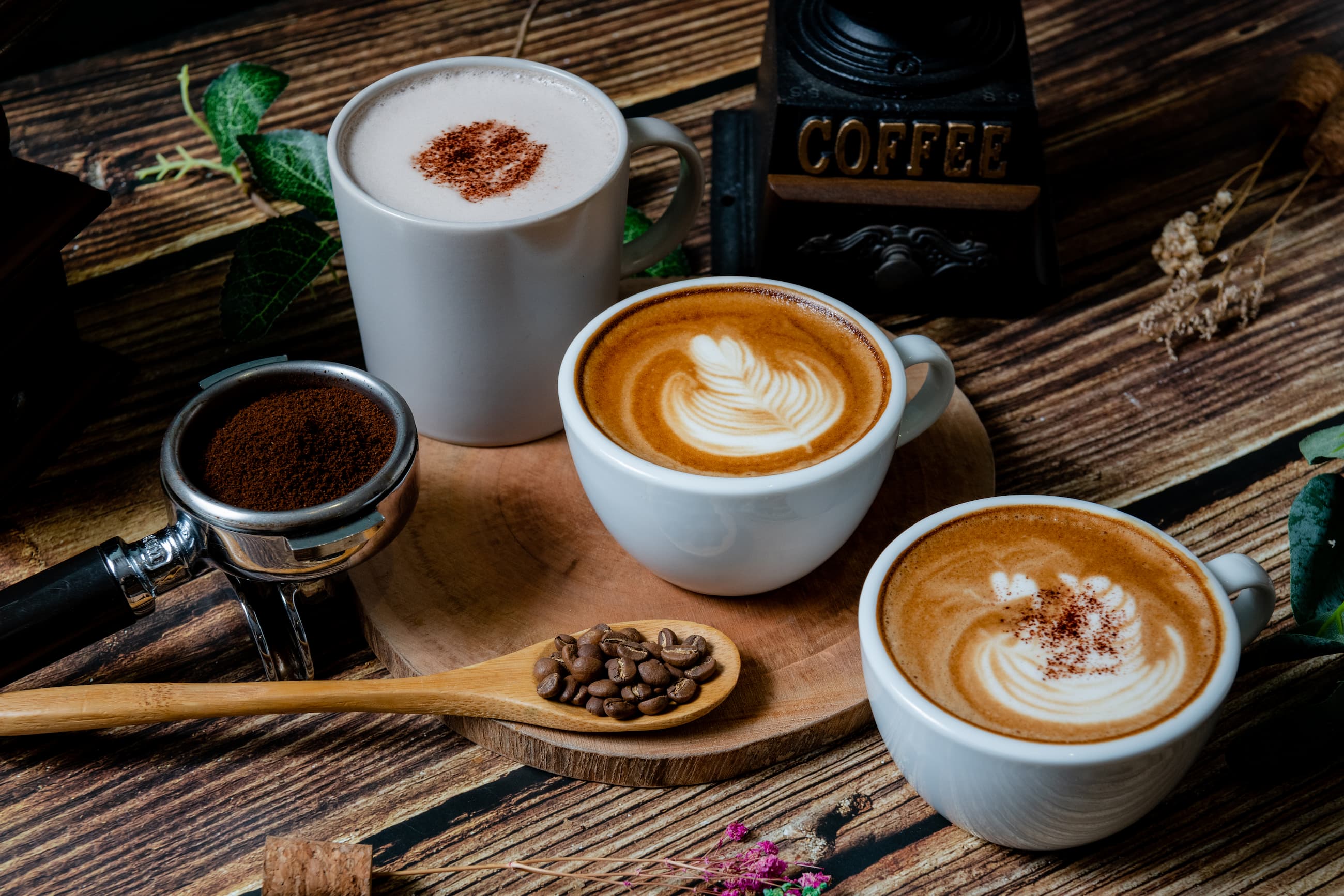 Drei Kaffeetassen mit Latte Art und Milchkaffee, dazu Siebträger mit gemahlenem Kaffee.