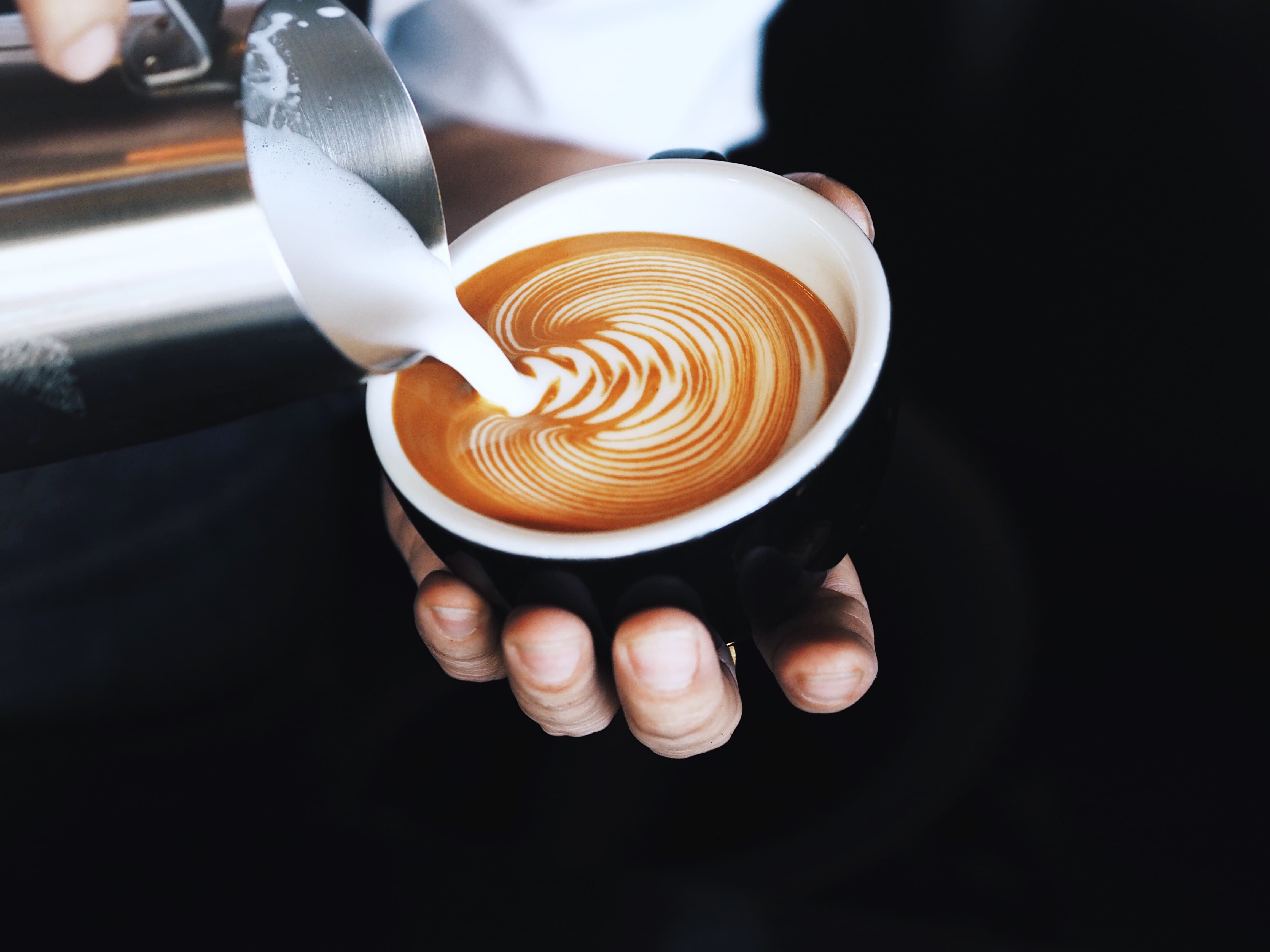 Barista gießt Milch in Espresso und zaubert Latte Art Muster in schwarzer Tasse.