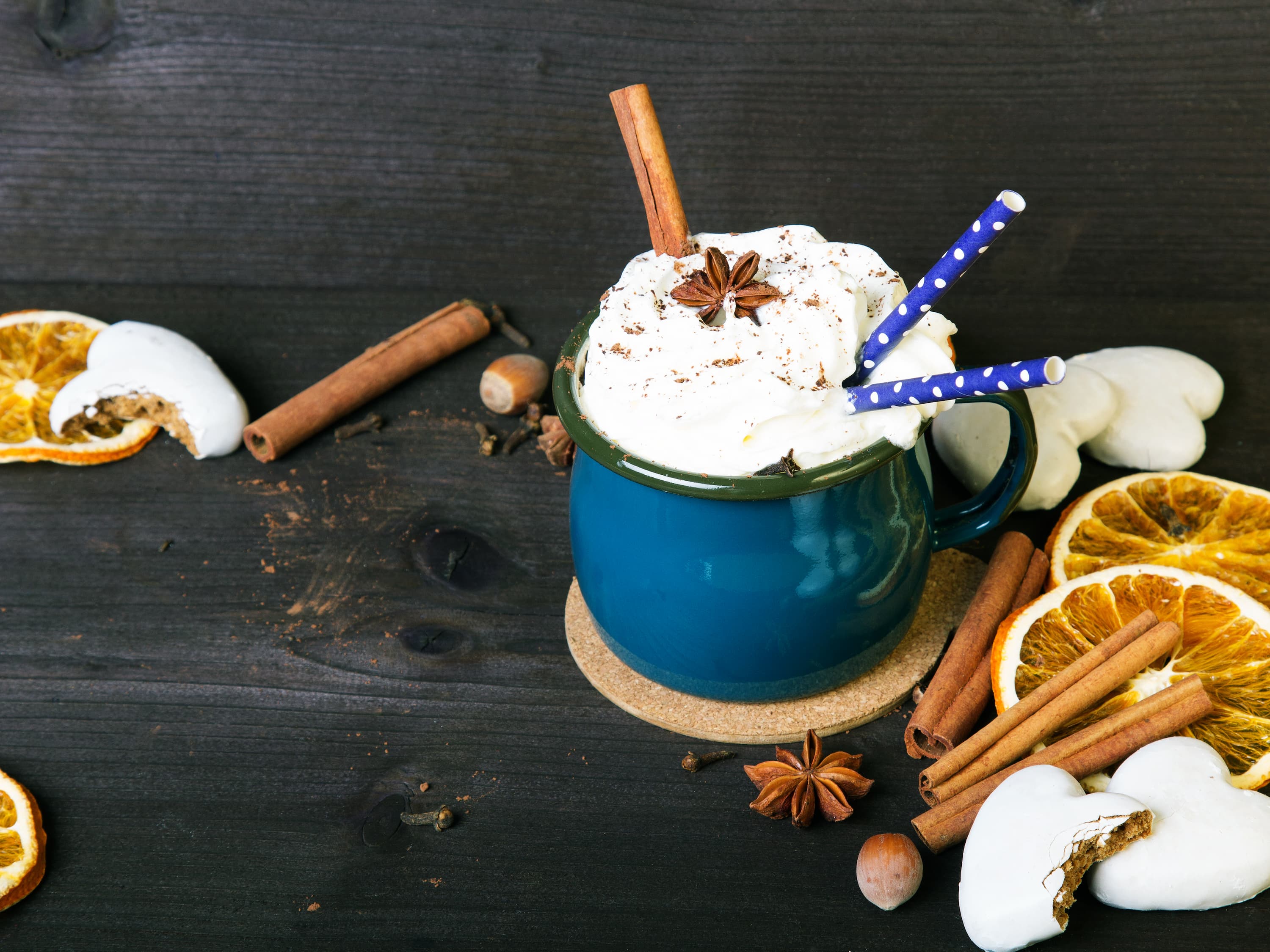 Tasse Kaffee mit Sahnehaube, Zimtstangen und getrockneten Orangenscheiben auf dunklem Holztisch.