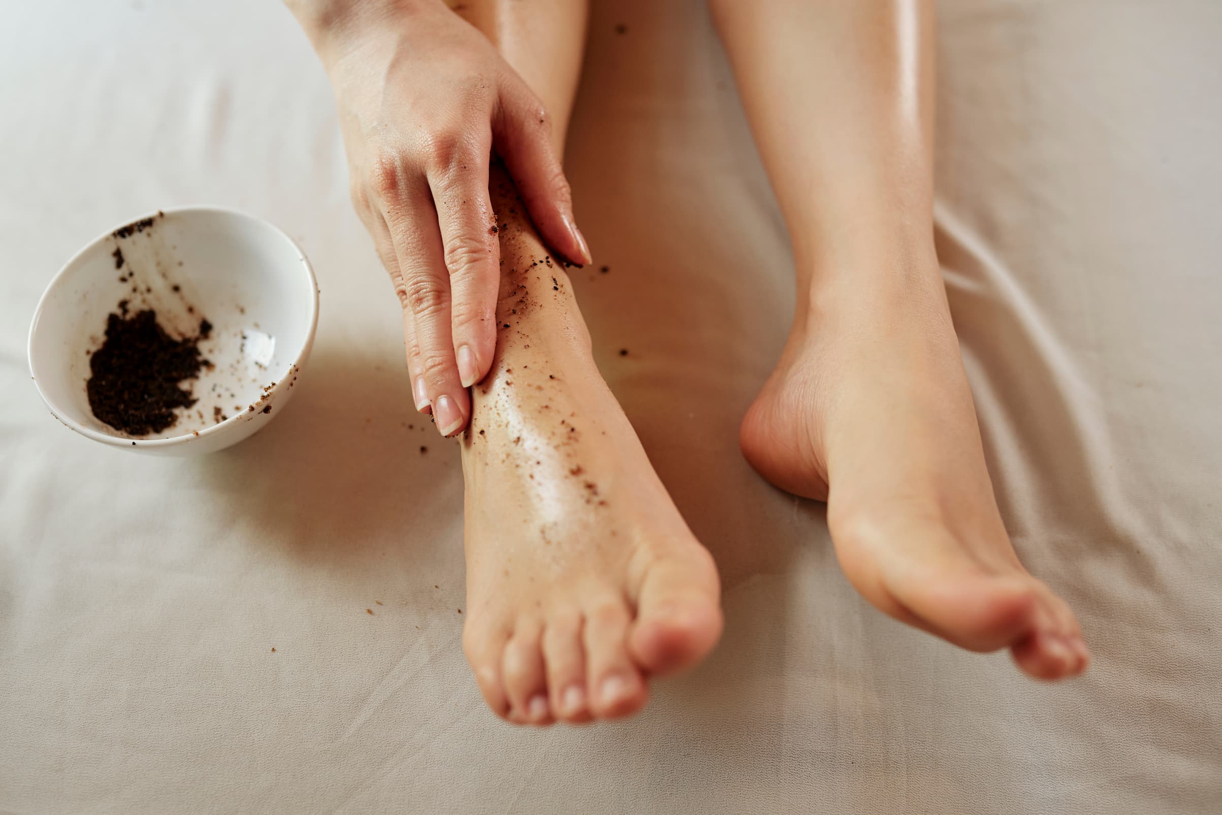Frau traegt ein natuerliches Kaffeepeeling auf ihre Beine auf, daneben steht eine Schale mit Kaffeepulver.