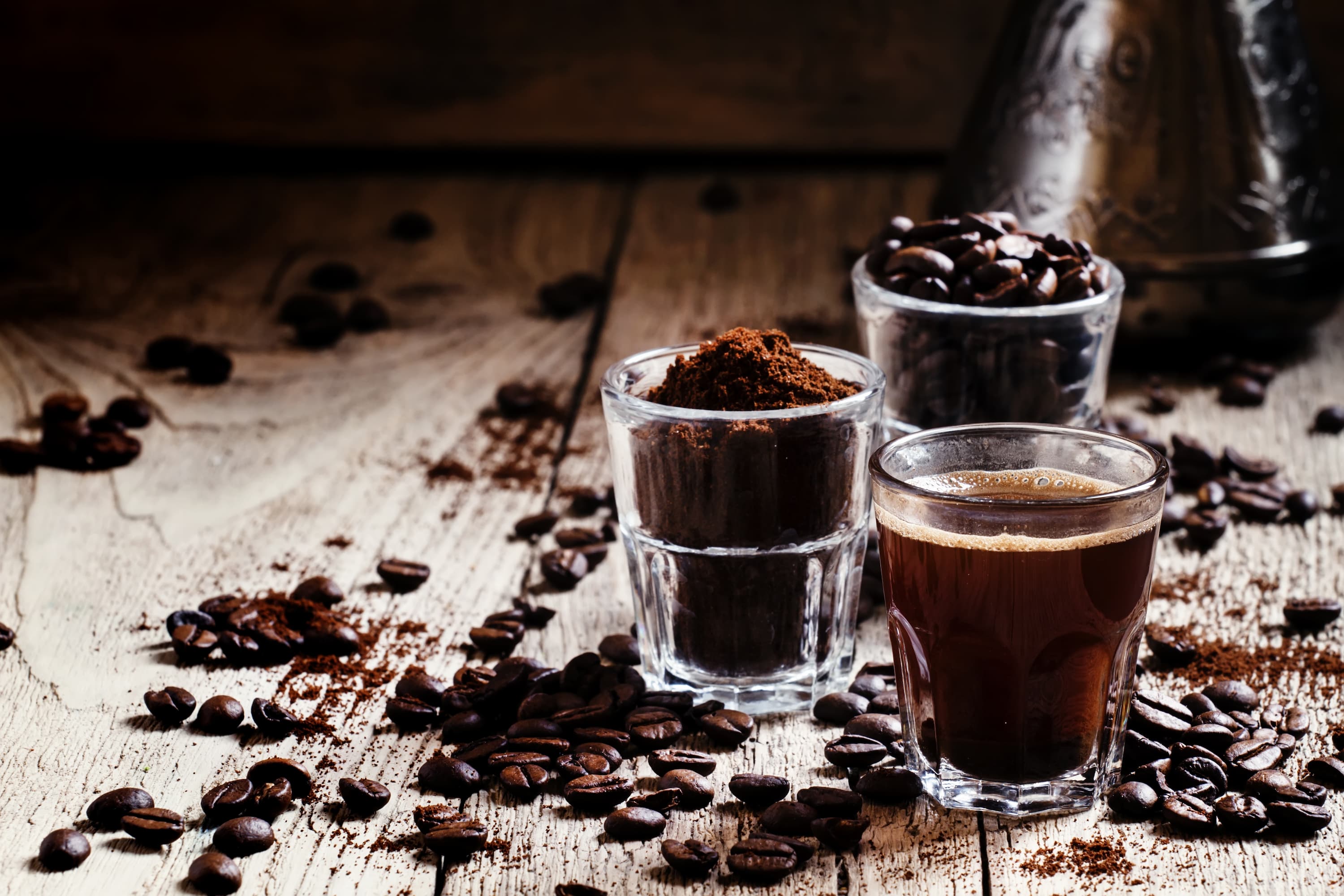 Drei Gläser mit Kaffeebohnen, Kaffeepulver und frischem Espresso auf Holztisch.
