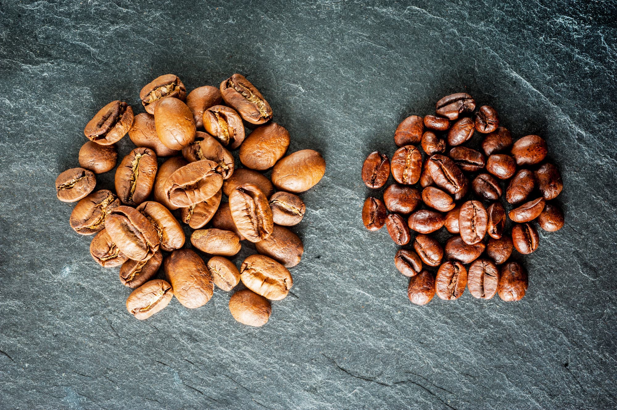 Zwei Häufchen Kaffeebohnen – links helle Röstung, rechts dunkle Röstung auf Steinplatte.