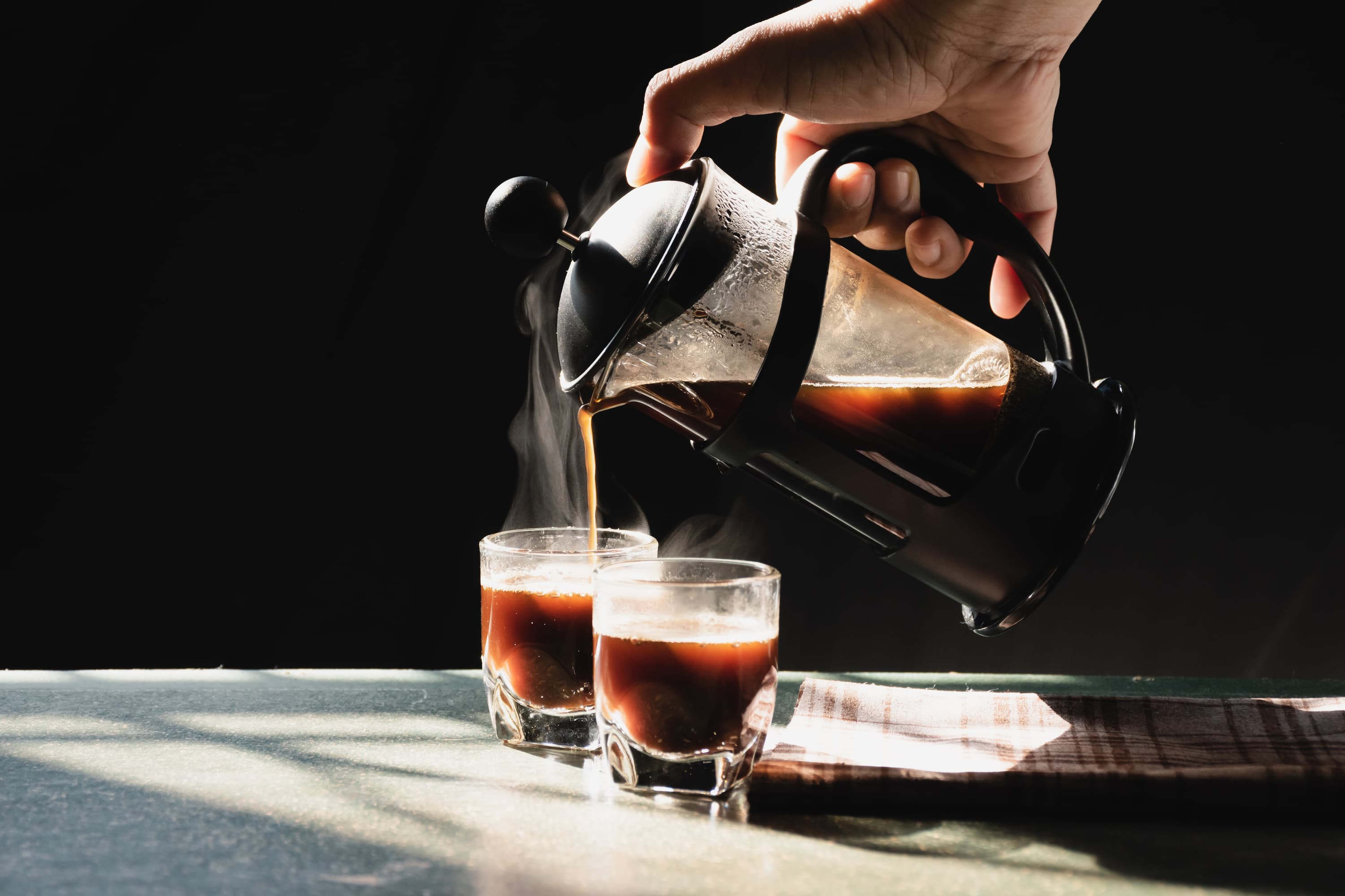 Hand gießt heißen Kaffee aus einer French Press in zwei Glaeser auf dunklem Hintergrund.