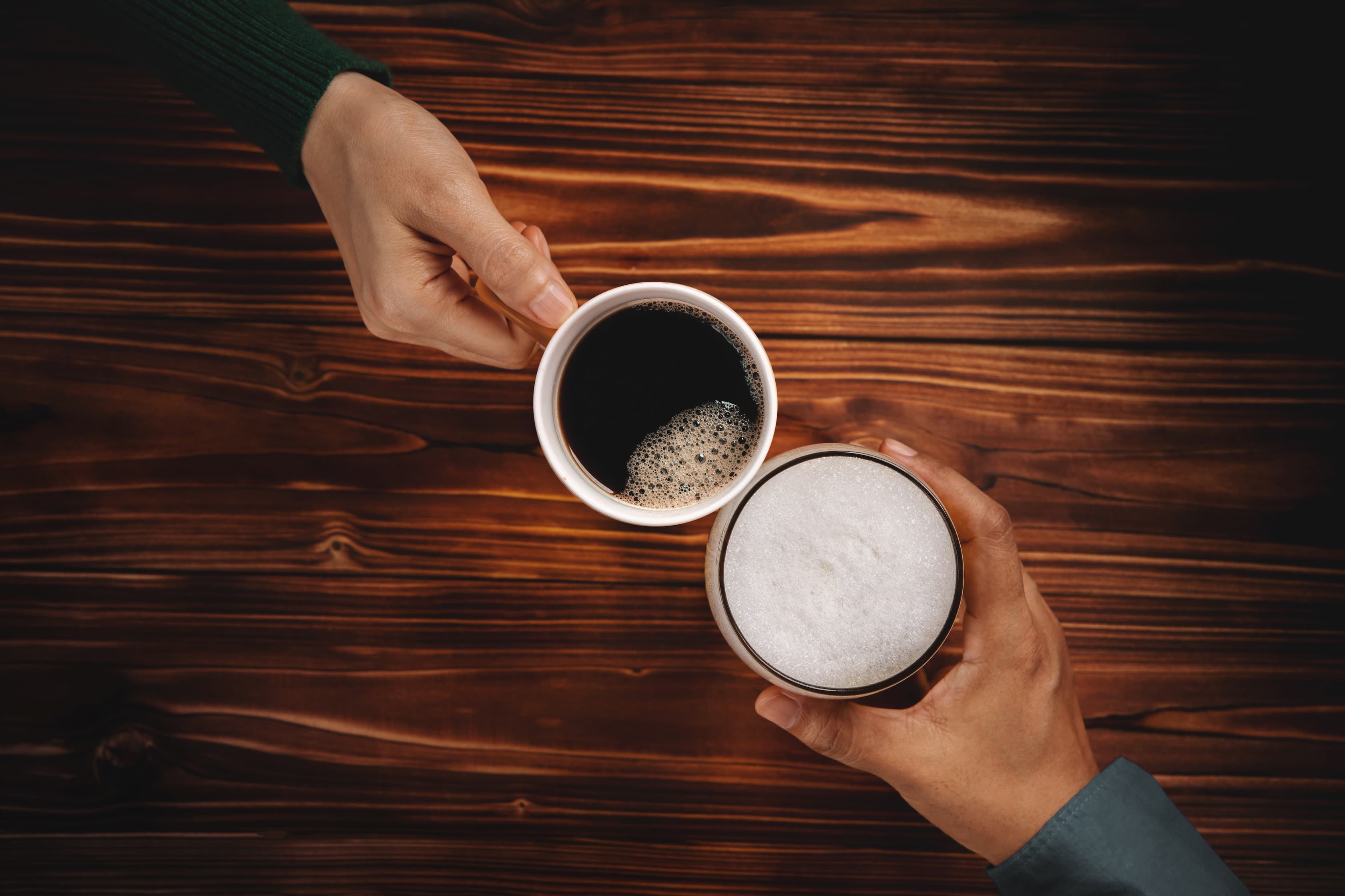 Zwei Hände halten Kaffeetassen mit schwarzem Kaffee und Cappuccino, Ansicht von oben.
